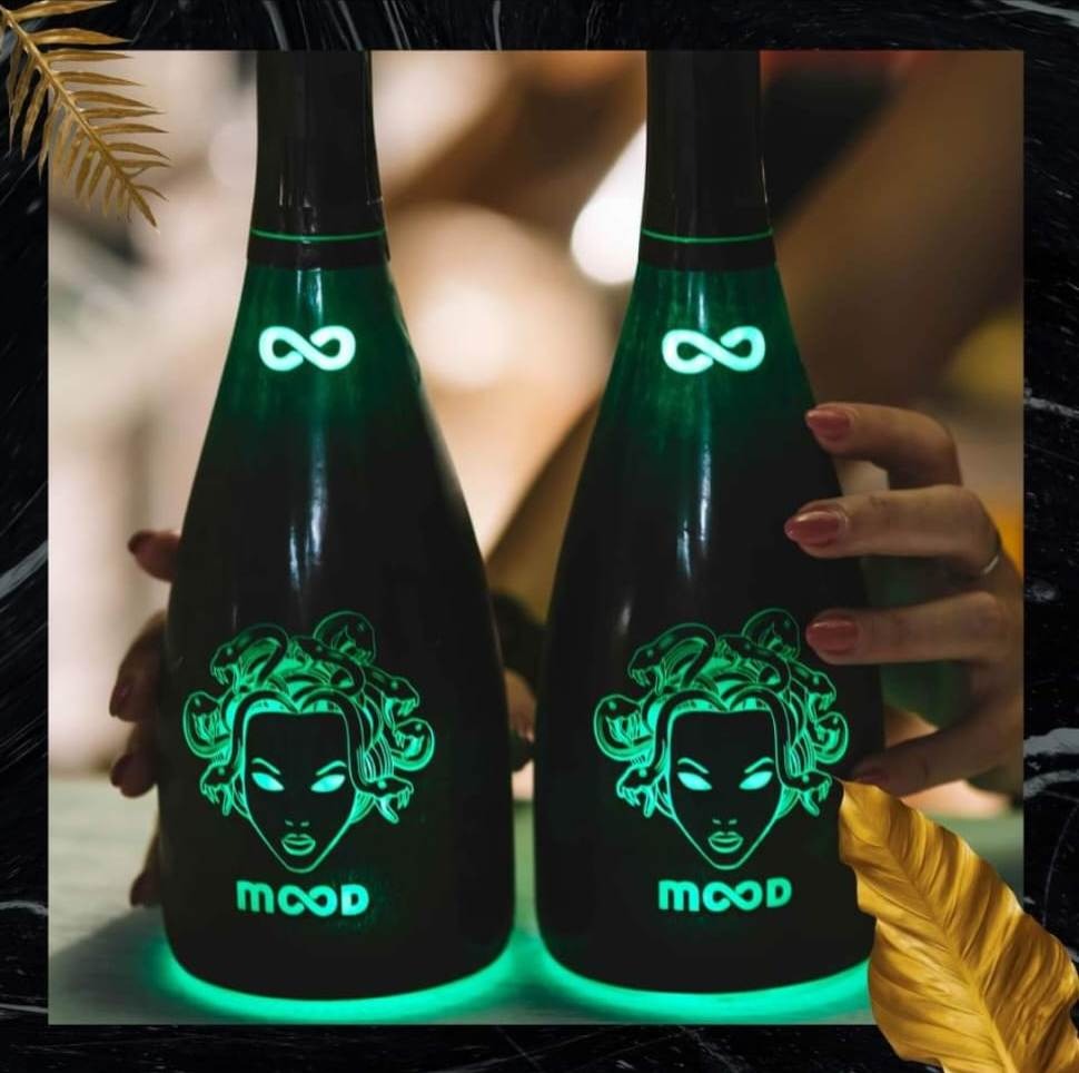 Dwie podświetlane butelki czarnego wina musującego MOOD z motywem Meduzy i symbolem nieskończoności, trzymane w dłoniach, na tle marmurowej tekstury.