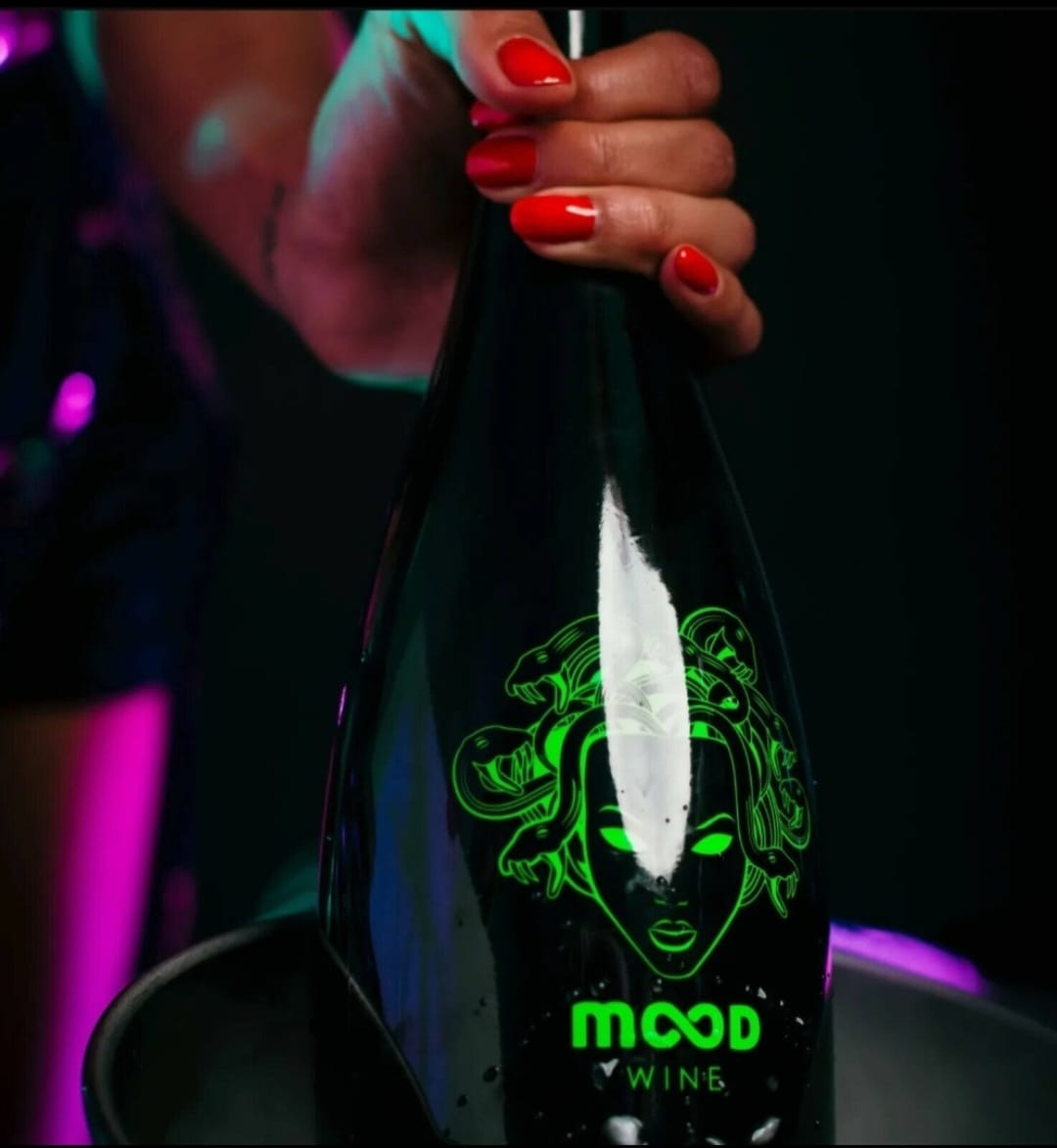 Czarna butelka wina z neonowo zielonym logo Meduzy trzymana przez dłoń z czerwonymi paznokciami, w tle fioletowe światło.