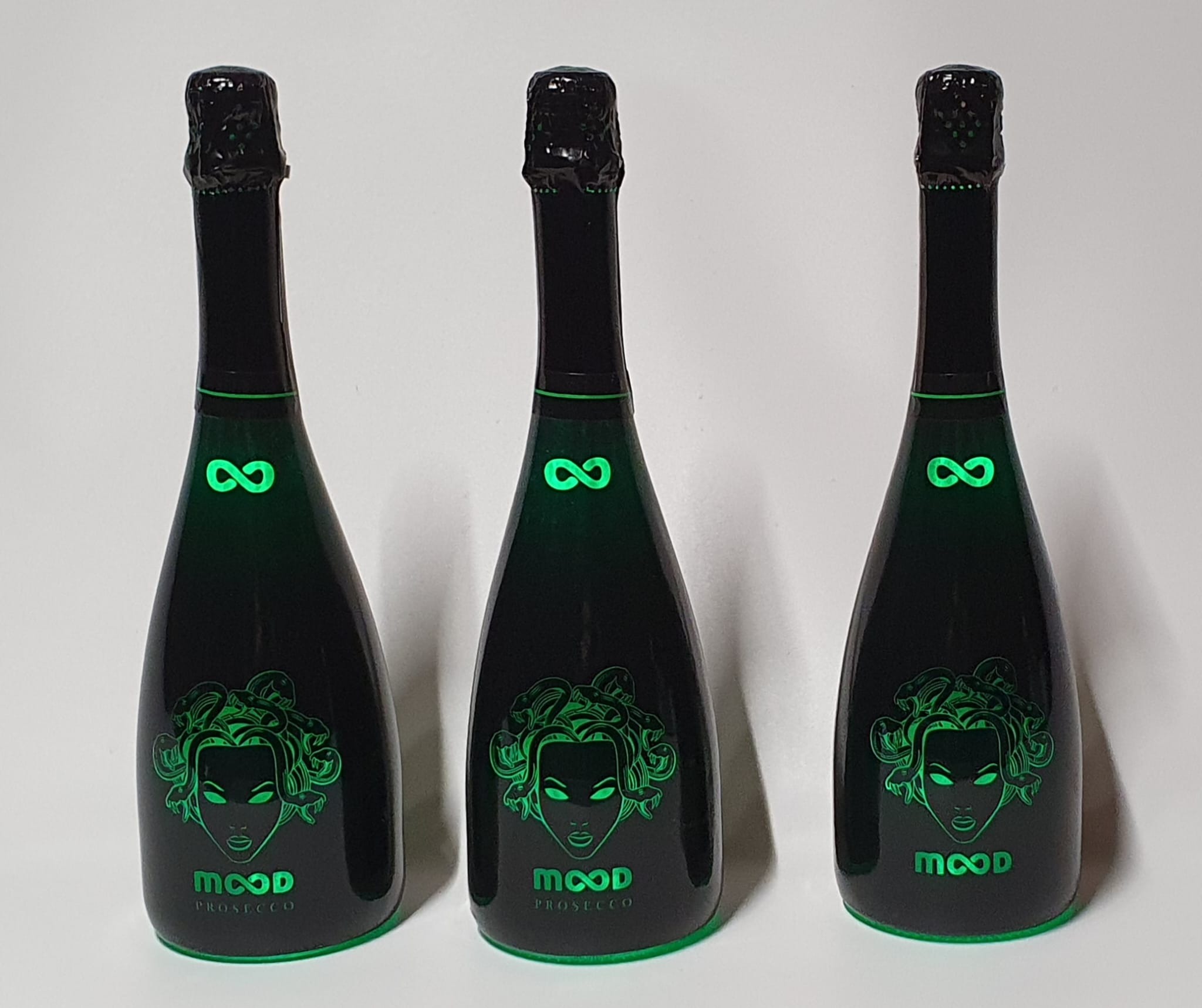 Trzy butelki czarnego prosecco z zielonym, świecącym wizerunkiem Meduzy i symbolem nieskończoności, ustawione obok siebie na białym tle.