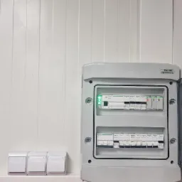 Rozdzielnica elektryczna NOARK z zainstalowanymi bezpiecznikami Schneider, zamontowana na białej ścianie z trzema włącznikami światła po lewej stronie, widok z bliska.