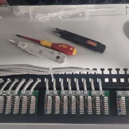 Panel krosowniczy z podłączonymi kablami sieciowymi, narzędzia (zaciskarka, nóż, śrubokręt Wiha) i niebieska taśma izolacyjna w szafie rack.