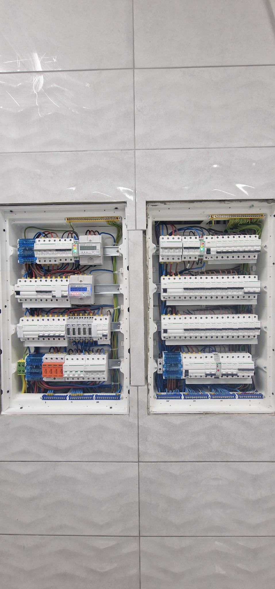 Dwa białe panele elektryczne z bezpiecznikami i przewodami, zamontowane na szarej ścianie z płytek o fakturze fal. W panelu po lewej stronie znajduje się licznik energii elektrycznej.