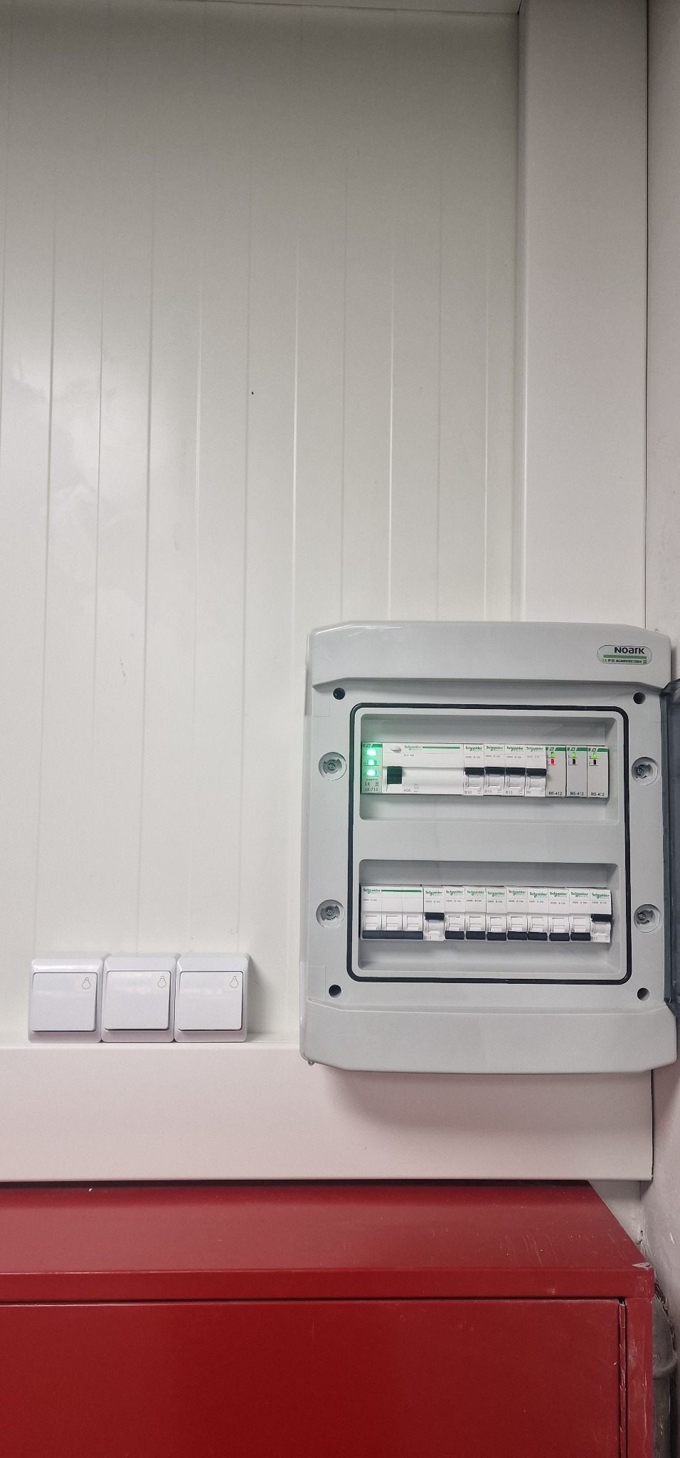 Rozdzielnica elektryczna NOARK z zainstalowanymi bezpiecznikami Schneider, zamontowana na białej ścianie z trzema włącznikami światła po lewej stronie, widok z bliska.