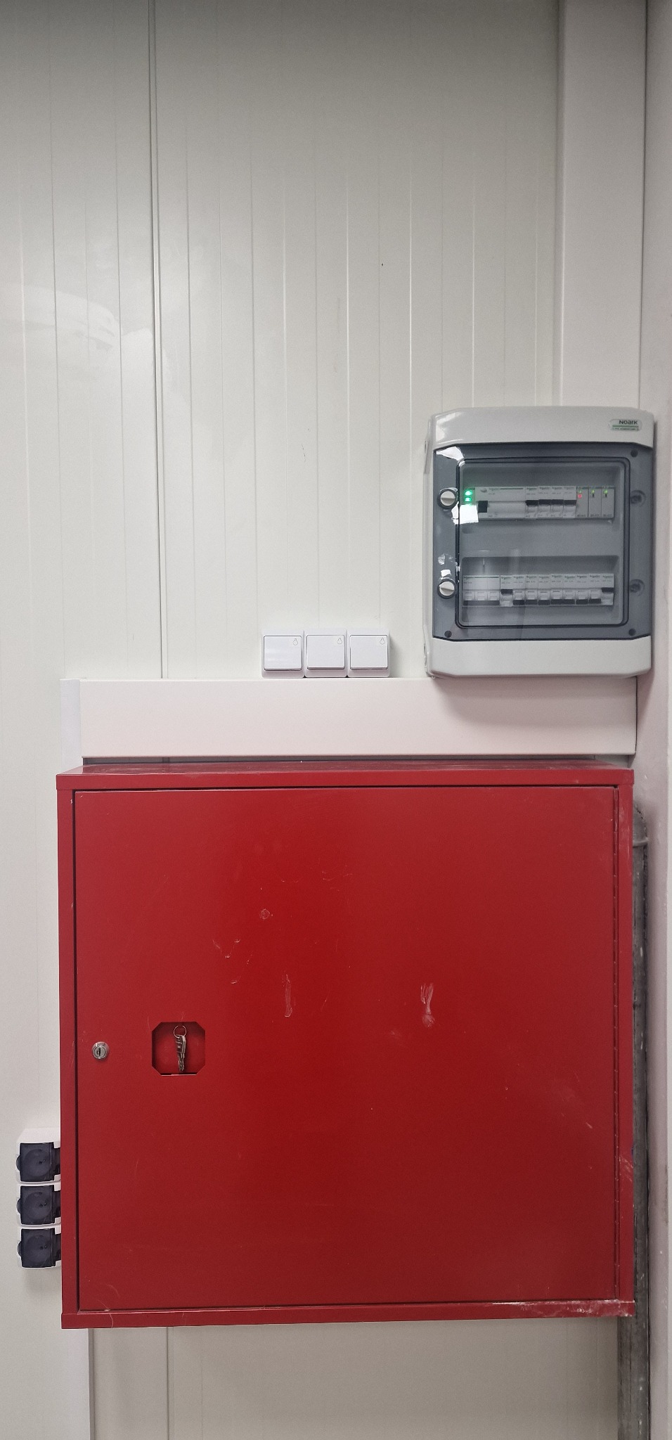 Zamknięta, czerwona szafka z kluczem w zamku, poniżej trzy gniazda elektryczne, nad nią trzy włączniki światła oraz skrzynka z bezpiecznikami z zieloną diodą LED.
