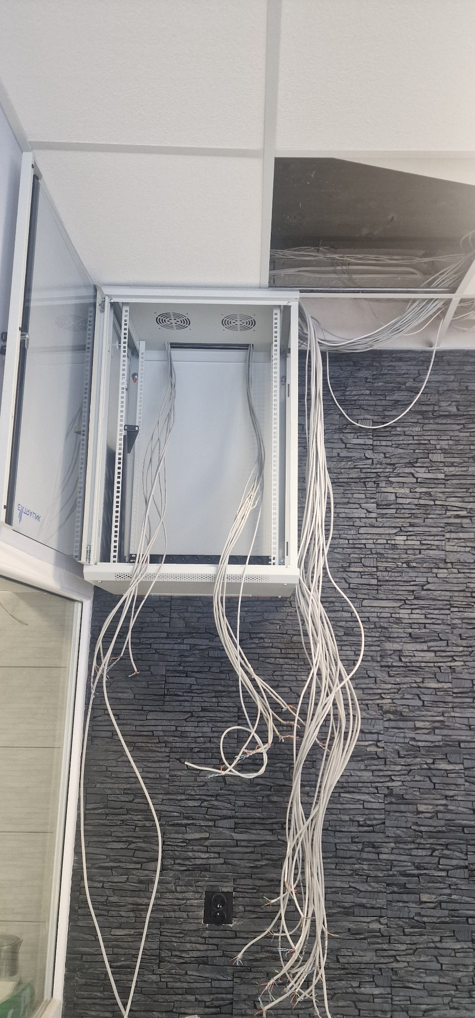 Szafa rack wisząca z otwartymi drzwiami, widoczne wentylatory i liczne przewody sieciowe zwisające w dół, ściana z szarej imitacji kamienia.
