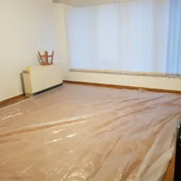 Pokój w Nowej Soli przygotowany do malowania: podłoga i listwy przypodłogowe zabezpieczone folią malarską, okno z żaluzjami pionowymi osłonięte folią, krzesło postawione na grzejniku.