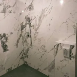 Wykończona kabina prysznicowa z marmurowymi płytkami, ciemna podłoga, bateria prysznicowa z deszczownicą i wnęka z instalacją elektryczną.