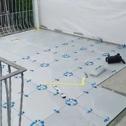 Układanie szarych płytek na balkonie, widoczne niebieskie klipsy poziomujące, poziomica i wiadro z narzędziami.