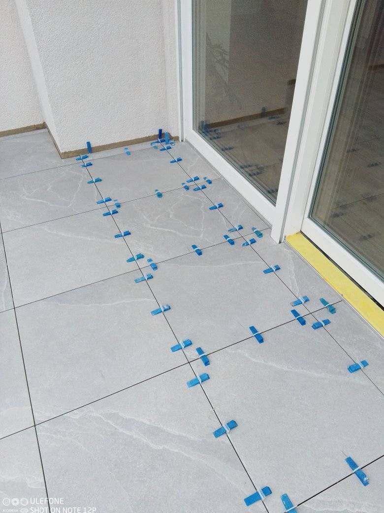 Układanie jasnoszarych płytek na podłodze balkonu z użyciem niebieskich krzyżyków dystansowych, widoczna ściana i przesuwne drzwi balkonowe.