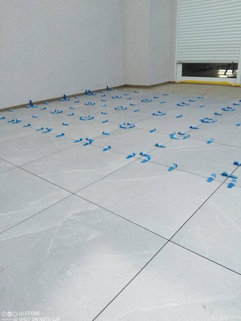 Jasnoszare płytki podłogowe ułożone w kwadraty, z niebieskimi klipsami poziomującymi spoiny, w trakcie prac wykończeniowych w pomieszczeniu z białymi ścianami i roletami.