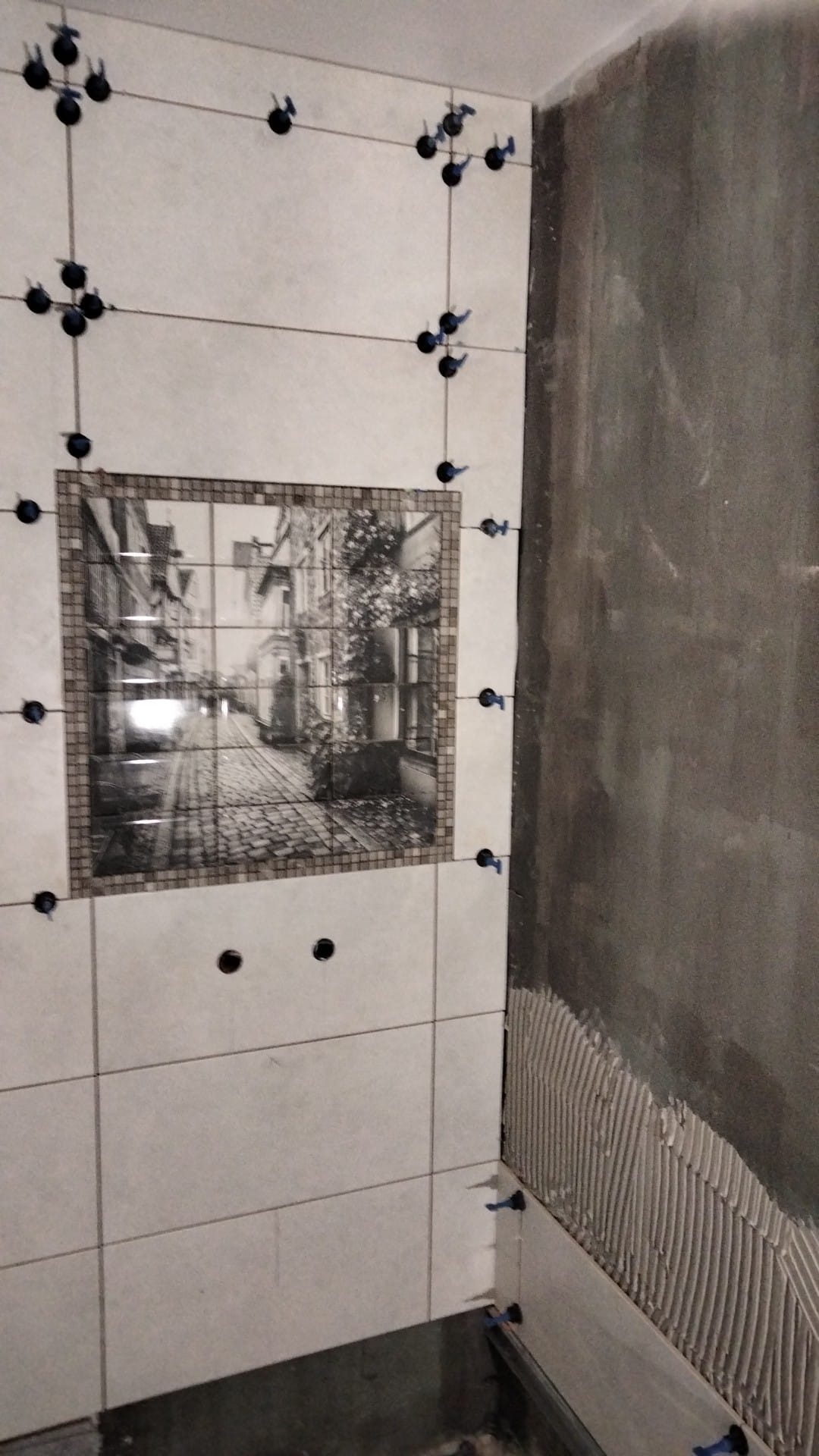 Układanie płytek z dekoracyjnym motywem uliczki w łazience, widoczne krzyżyki dystansowe i fragment ściany z nałożonym klejem.