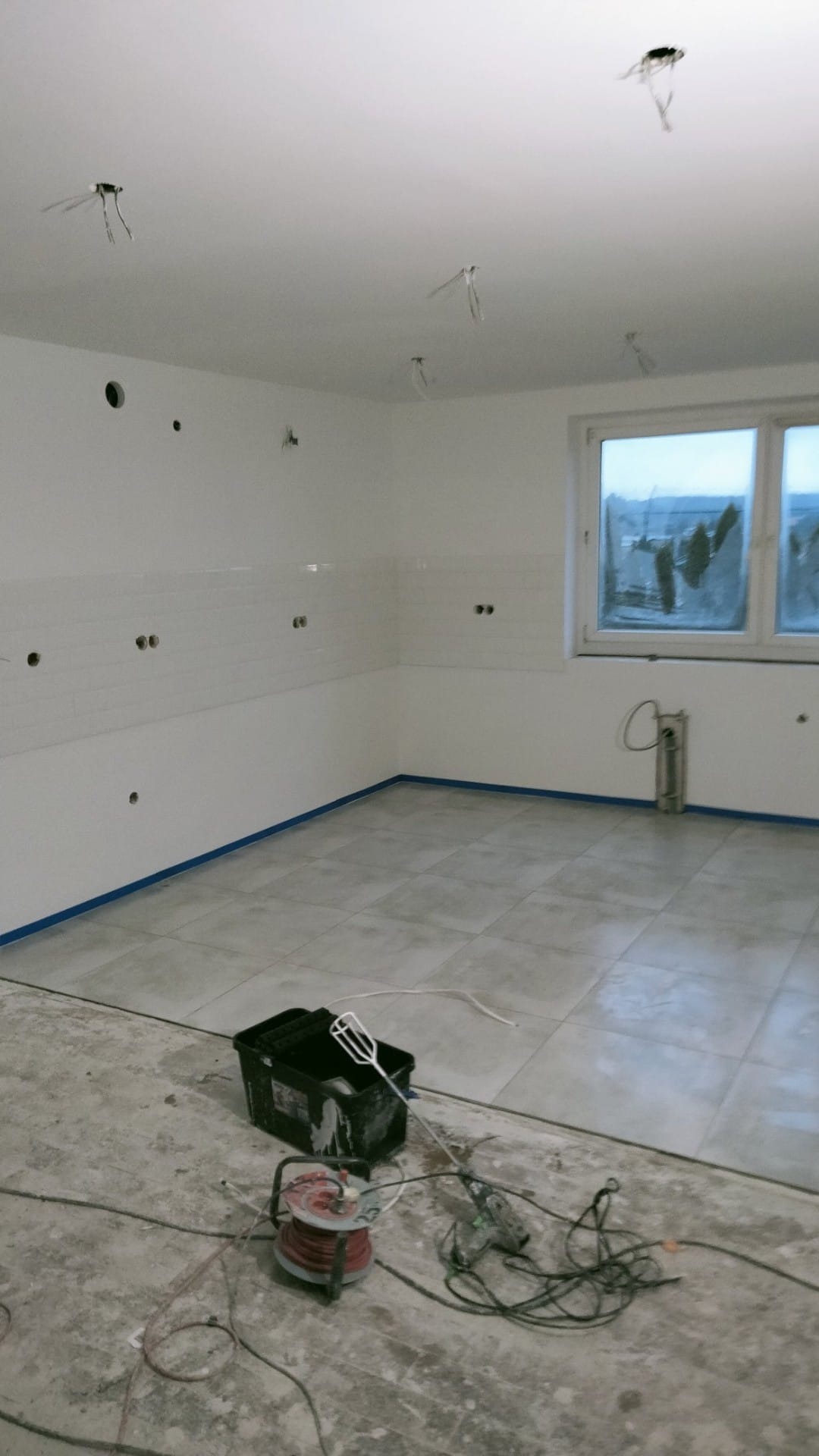 Wnętrze w trakcie prac wykończeniowych: ułożone szare płytki podłogowe, białe ściany z częściowo położonymi białymi płytkami, zabezpieczone niebieską taśmą, widoczne wystające kable i puszki...