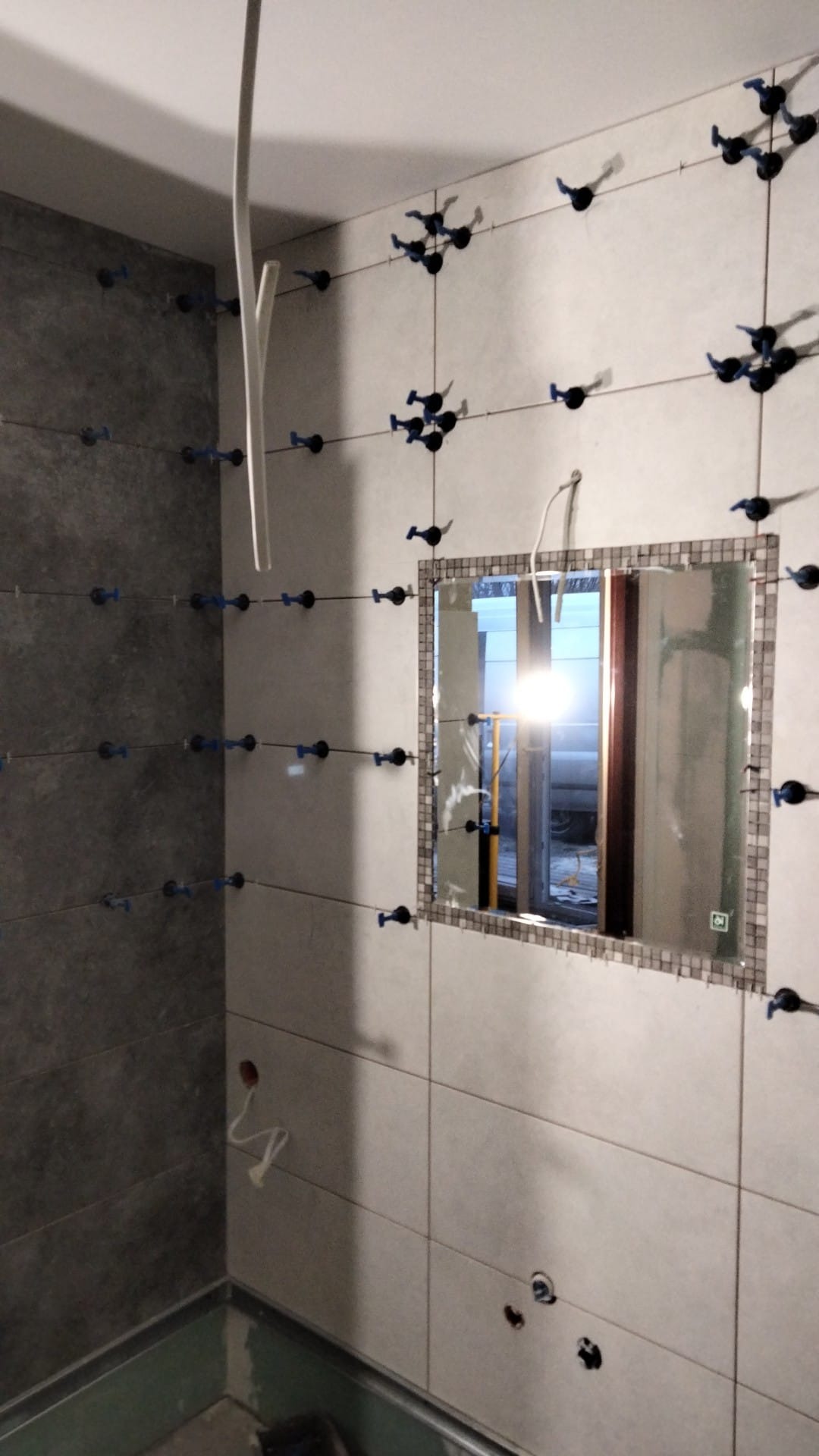 Ściana łazienki w trakcie układania płytek, z widocznym lustrem w mozaikowej ramie i systemem poziomowania płytek.