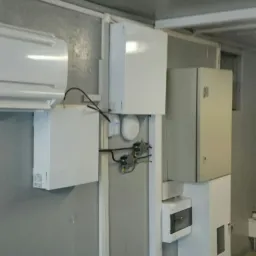 Wewnętrzna instalacja elektryczna z licznymi białymi skrzynkami, przewodami i czujnikiem ruchu na szarej ścianie w pomieszczeniu.