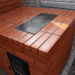 Murowany piec z czerwonej cegły z żeliwnymi drzwiczkami i płytą grzewczą, ustawiony na zewnątrz przy betonowym ogrodzeniu.
