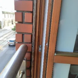 Widok z okna balkonowego z drewnianą ramą, z widoczną ceglaną ścianą i fragmentem metalowej balustrady, na tle ulicy z budynkami i samochodem.