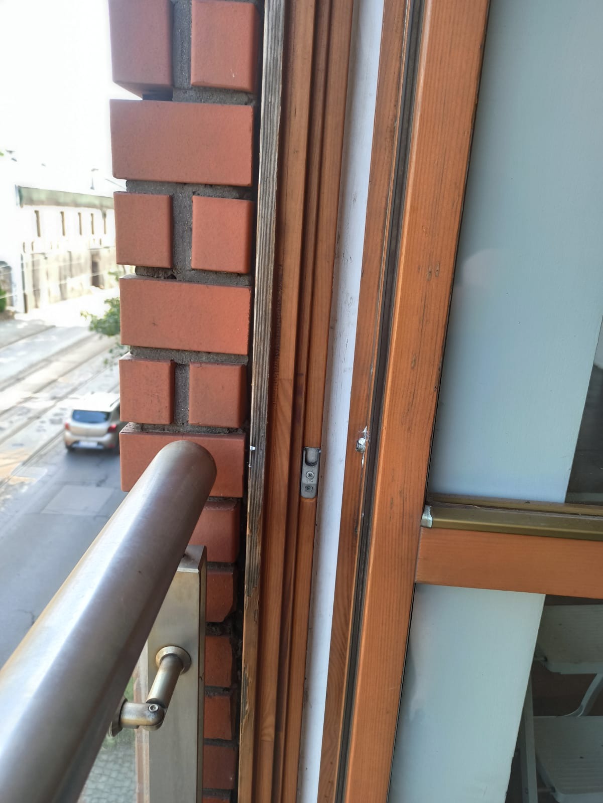 Widok z okna balkonowego z drewnianą ramą, z widoczną ceglaną ścianą i fragmentem metalowej balustrady, na tle ulicy z budynkami i samochodem.