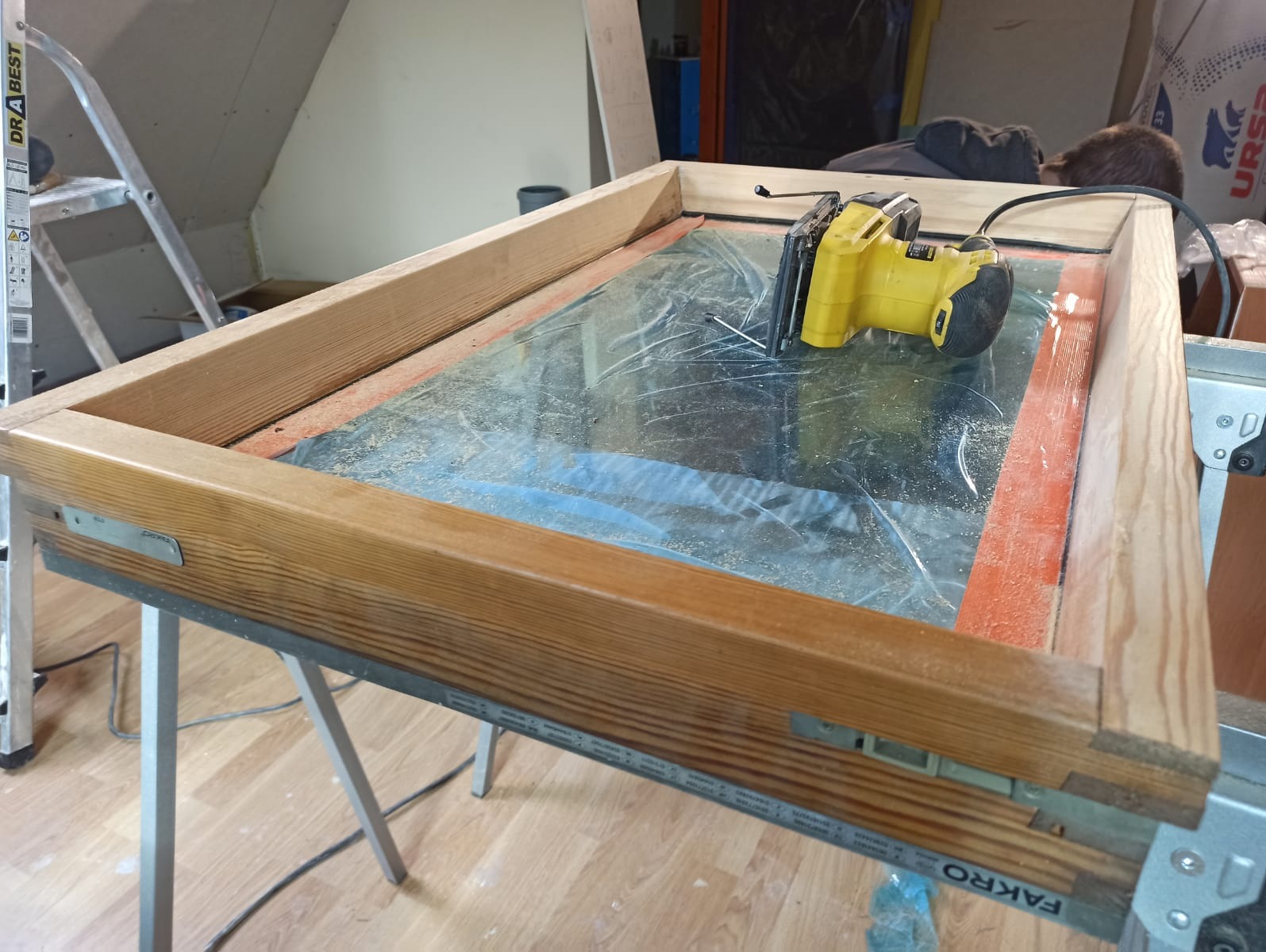 Żółta piła elektryczna na drewnianej ramie okiennej z szybą, ustawionej na metalowym stole warsztatowym, w tle drabina i nieporządek.