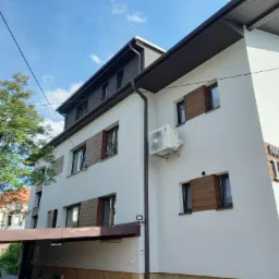 Biały, trzypiętrowy budynek z brązowymi okiennicami, klimatyzacją na ścianie i numerem 15 'Na Banie' na fasadzie, widziany z dołu pod kątem.