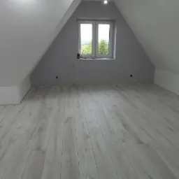 Poddasze z oknem i panelami podłogowymi w odcieniach szarości, widoczne niedokończone instalacje oświetleniowe w suficie.