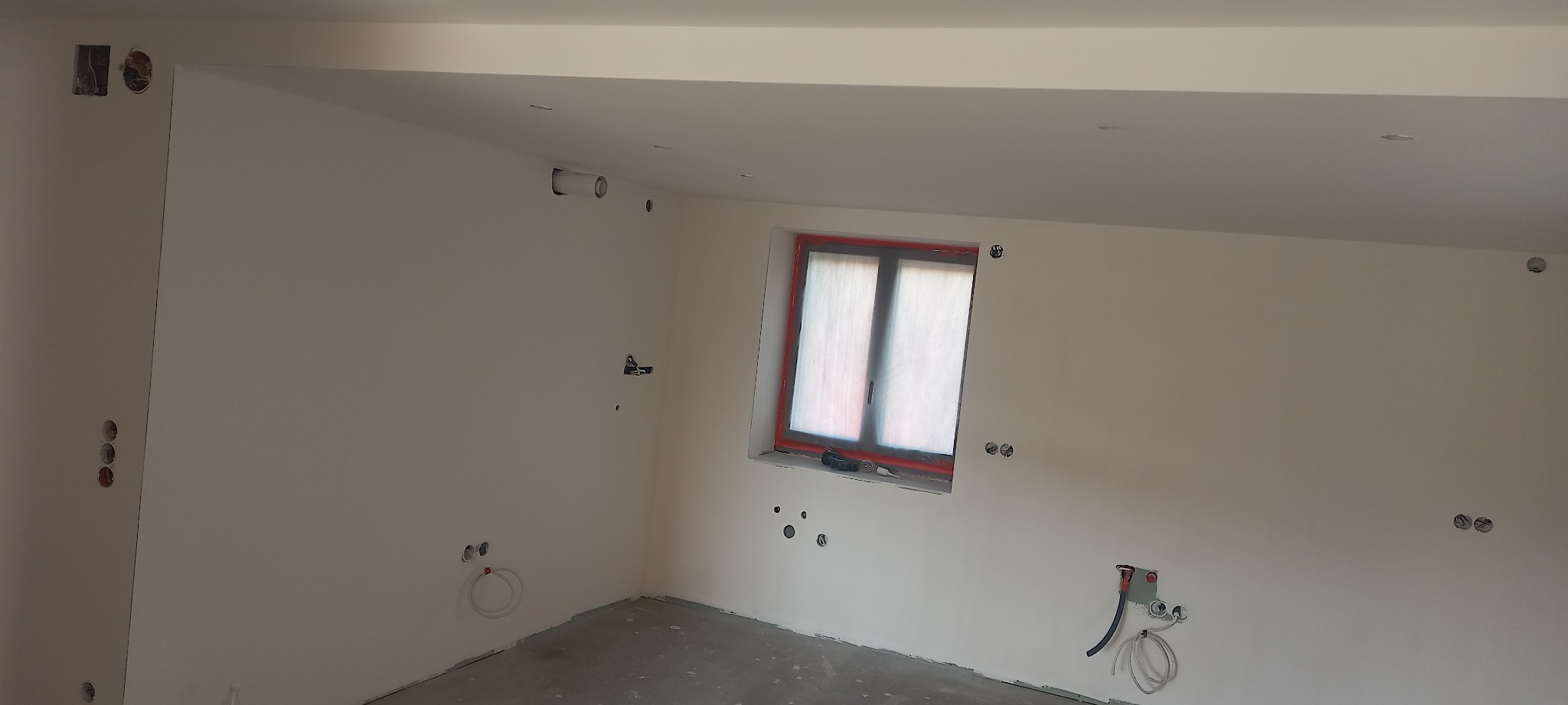 Wnętrze w trakcie prac wykończeniowych: świeżo pomalowane ściany na biało, widoczne punkty elektryczne i hydrauliczne, okno zabezpieczone folią malarską, surowa podłoga.