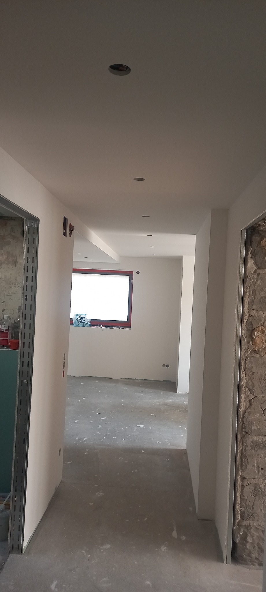 Korytarz w trakcie remontu z widocznymi nowymi ścianami z płyt karton gips, przygotowany do malowania, z surową podłogą i oknem w czerwonej ramie.