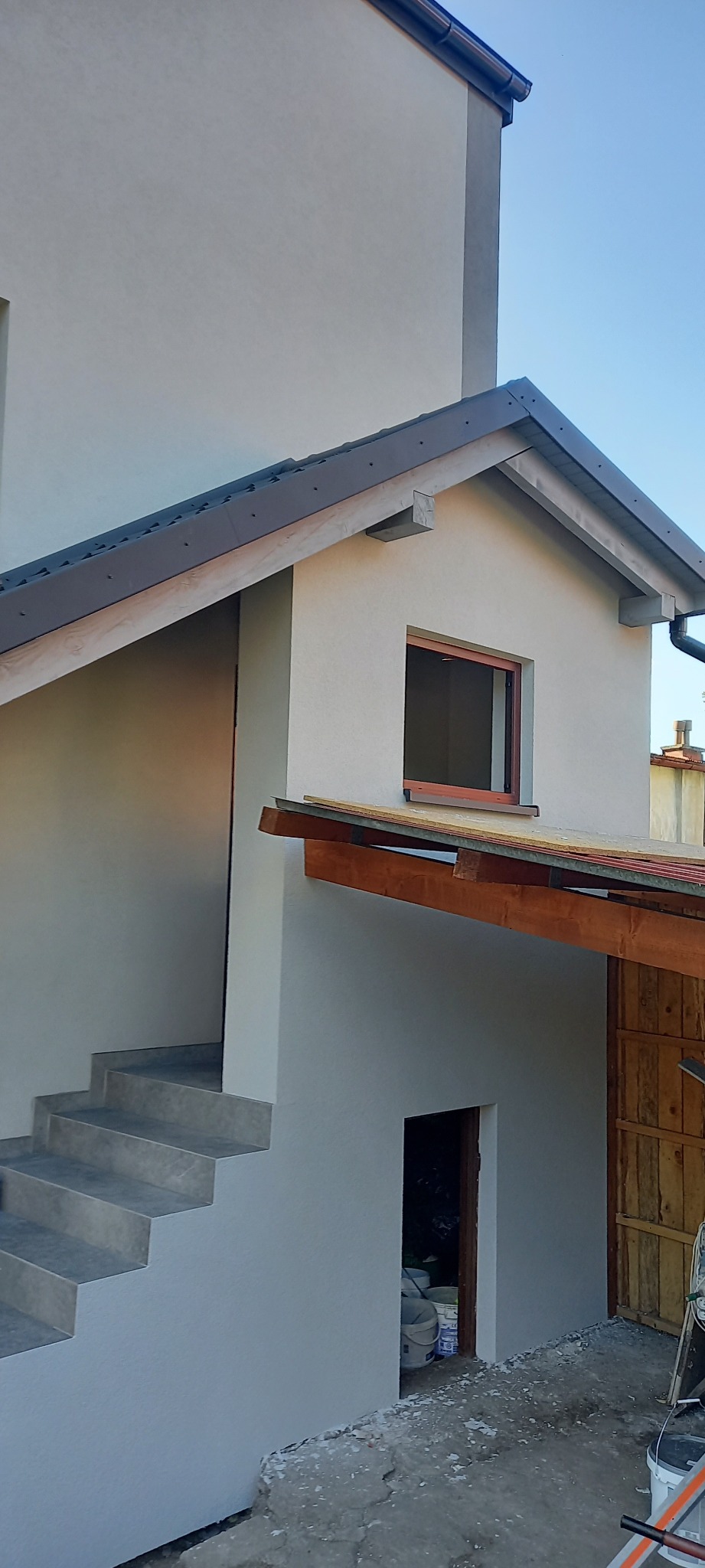 Elewacja budynku po remoncie z widocznymi schodami zewnętrznymi, oknem w poddaszu i nowym orynnowaniem. Widoczne wiaderka po farbach i inne ślady prac budowlanych.