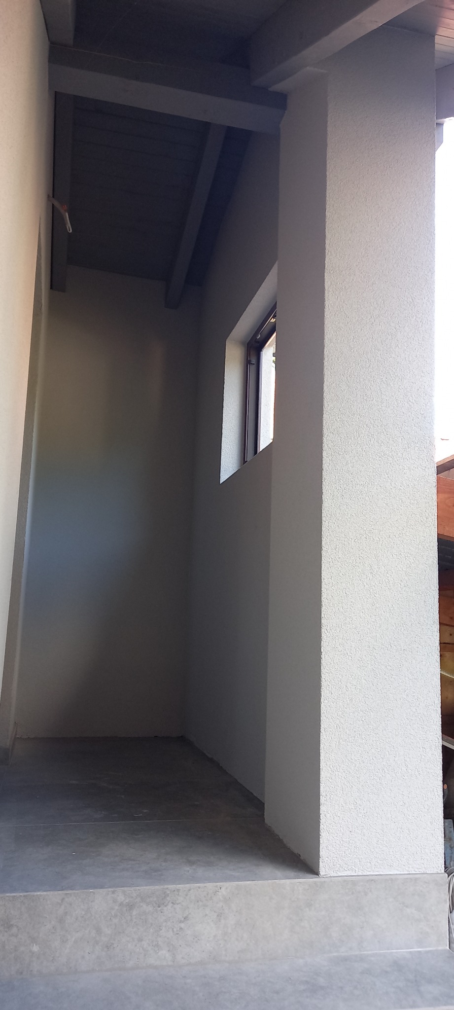 Pomalowana na szaro wnęka z oknem i betonową podłogą, widoczne elementy konstrukcji drewnianej pod dachem.