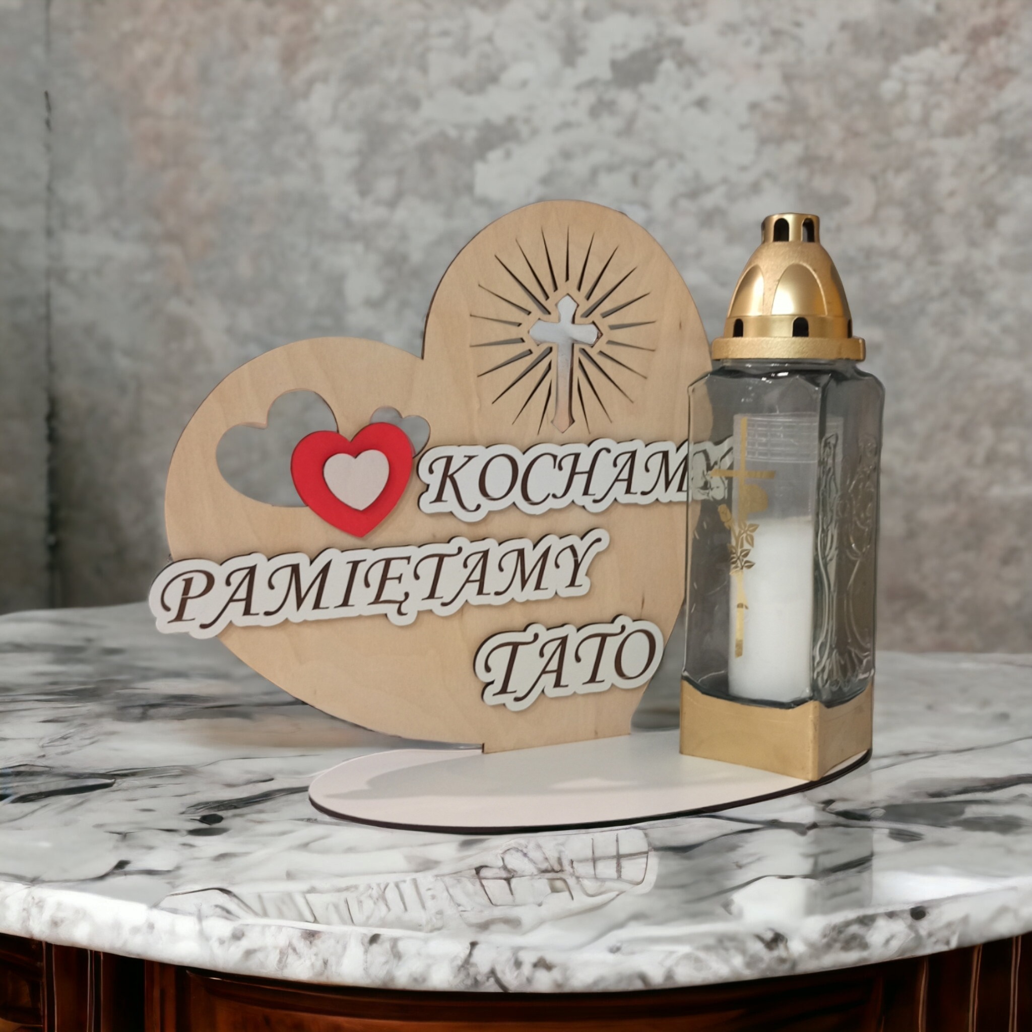 Drewniana ozdoba w kształcie serca z napisem 'Kochamy Pamiętamy Tato' oraz znicz w szklanej obudowie z złotymi elementami, stojące na marmurowym blacie.