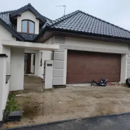 Nowoczesny dom jednorodzinny w Laskach z brązową bramą garażową i czarną dachówką, widok na elewację frontową w pochmurny dzień.