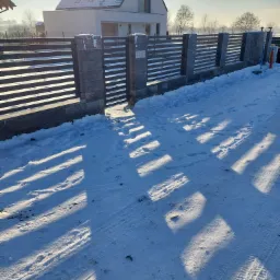 Nowoczesne ogrodzenie panelowe z szarego betonu łupanego i poziomych, ciemnych przęseł, brama wjazdowa, zimowy krajobraz z ośnieżonym podjazdem i domem w tle, ostre cienie rzucane przez słońce.