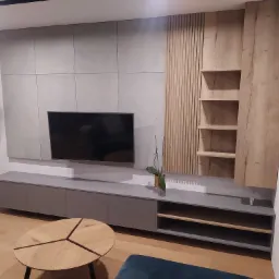 Nowoczesny salon z szarą ścianą z paneli, szafką RTV, drewnianymi elementami dekoracyjnymi i okrągłym stolikiem kawowym z drewnianym blatem.