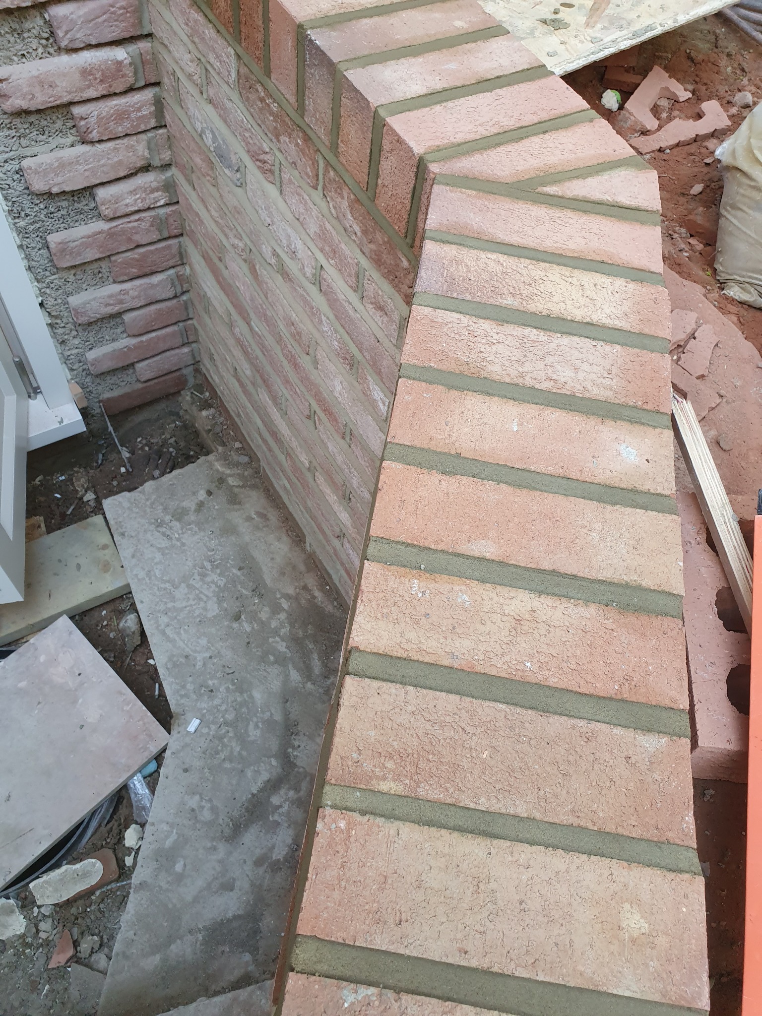 Murowana ściana z cegły klinkierowej w trakcie budowy, widoczne spoiny cementowe, fragment okna i gruz budowlany w tle.