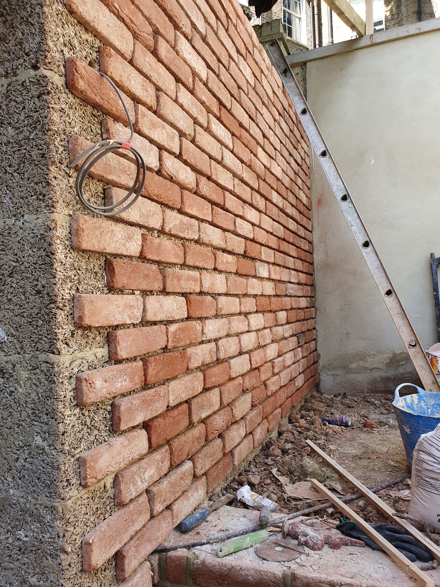 Fragment ściany zewnętrznej z ociepleniem i nową okładziną z cegły klinkierowej w trakcie montażu, widoczne narzędzia i materiały budowlane, drabina oparta o ścianę sąsiedniego budynku.