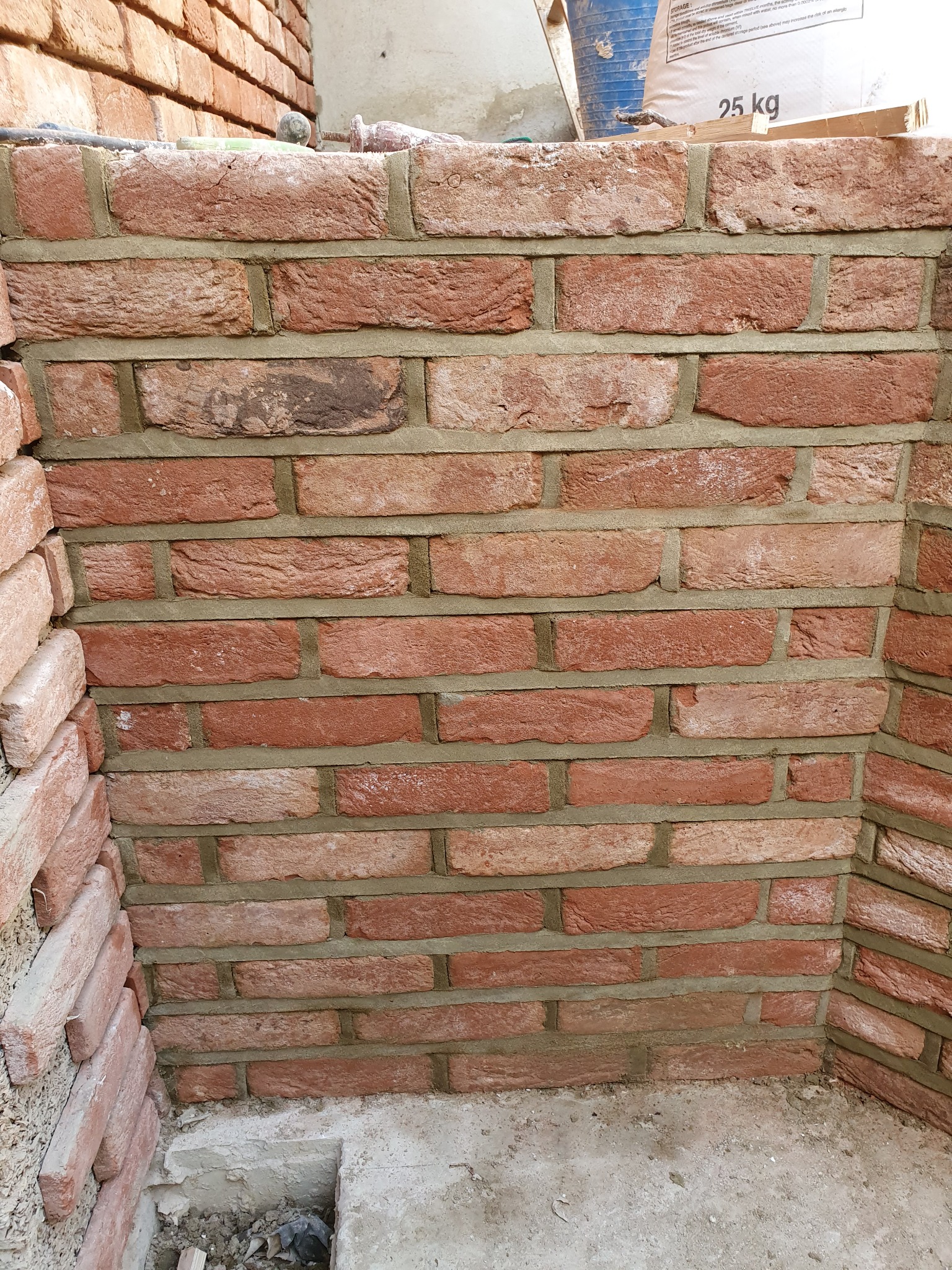 Świeżo wymurowana ściana z czerwonej cegły klinkierowej z widoczną zaprawą murarską, fragment podłogi betonowej z otworem w rogu.