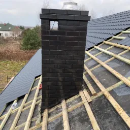 Montaż grafitowej dachówki na drewnianej konstrukcji z widocznym czarnym, ceglanym kominem z otworem wentylacyjnym i fragmentem krajobrazu w tle.