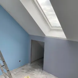 Wnętrze pokoju na poddaszu z pomalowanymi ścianami na niebiesko i szaro, widoczna drabina aluminiowa i okno dachowe.