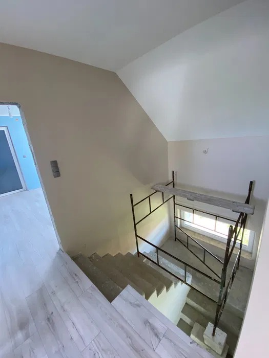 Wnętrze domu w trakcie remontu: widok na schody z surowego betonu, częściowo zabezpieczone rusztowaniem, z fragmentem pomalowanej ściany w odcieniach beżu i ułożoną podłogą z paneli imitujących...