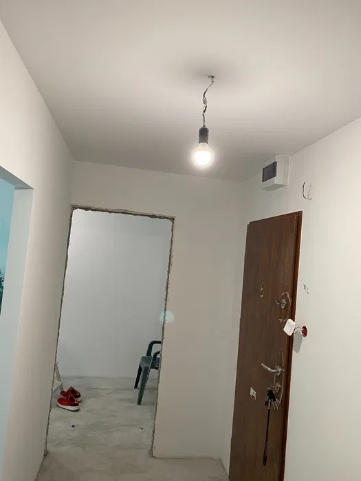 Remont korytarza w Toruniu: widok na surowy otwór drzwiowy, gołe ściany przygotowane do malowania, prowizoryczna żarówka zwisająca z sufitu, czerwone buty i krzesło w tle.