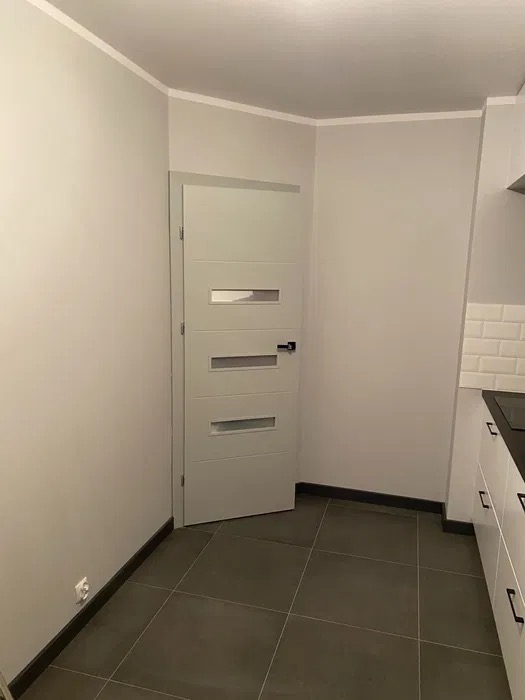 Nowoczesna szara łazienka z drzwiami z przeszkleniami i ciemną, kwadratową glazurą podłogową, widok na fragment zabudowy kuchennej z białymi szafkami i czarnym blatem.