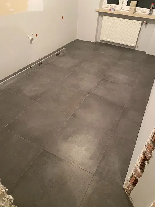 Ułożone ciemnoszare płytki podłogowe w pomieszczeniu z grzejnikiem, poziomica przy ścianie, ślady po instalacji na ścianie.