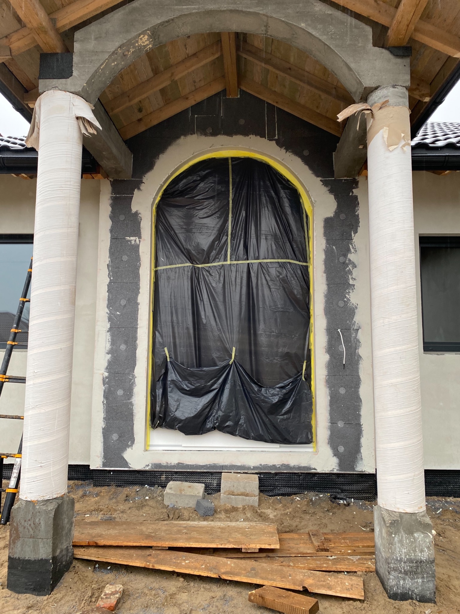 Elewacja domu w trakcie ocieplania: styropian na ścianie, okno zabezpieczone folią, kolumny owinięte folią ochronną, widoczne rusztowanie w tle.