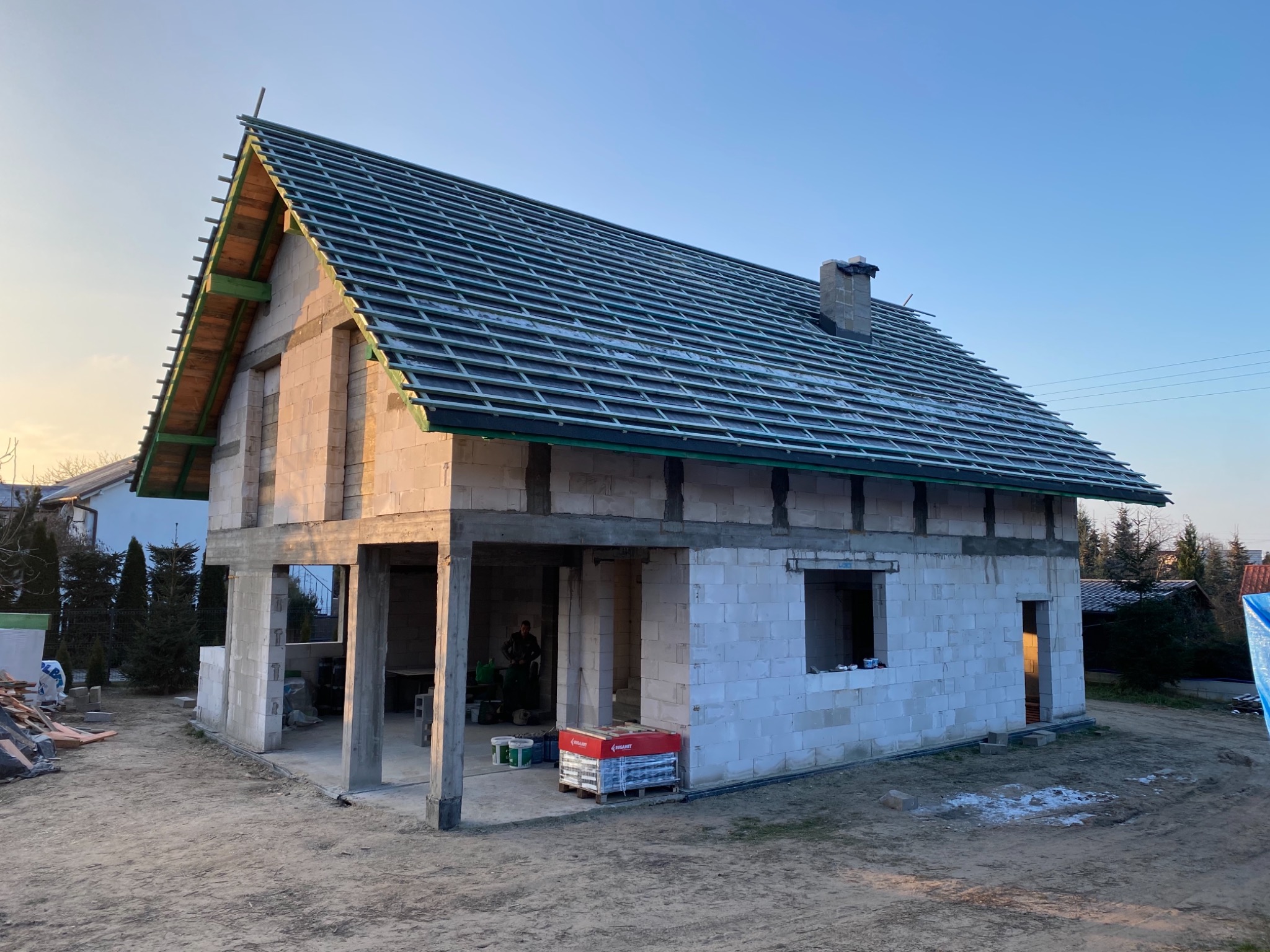 Budowa domu jednorodzinnego w trakcie realizacji: ściany z betonu komórkowego, drewniana konstrukcja dachu przygotowana pod pokrycie, widoczne materiały budowlane i osoba wewnątrz budynku.
