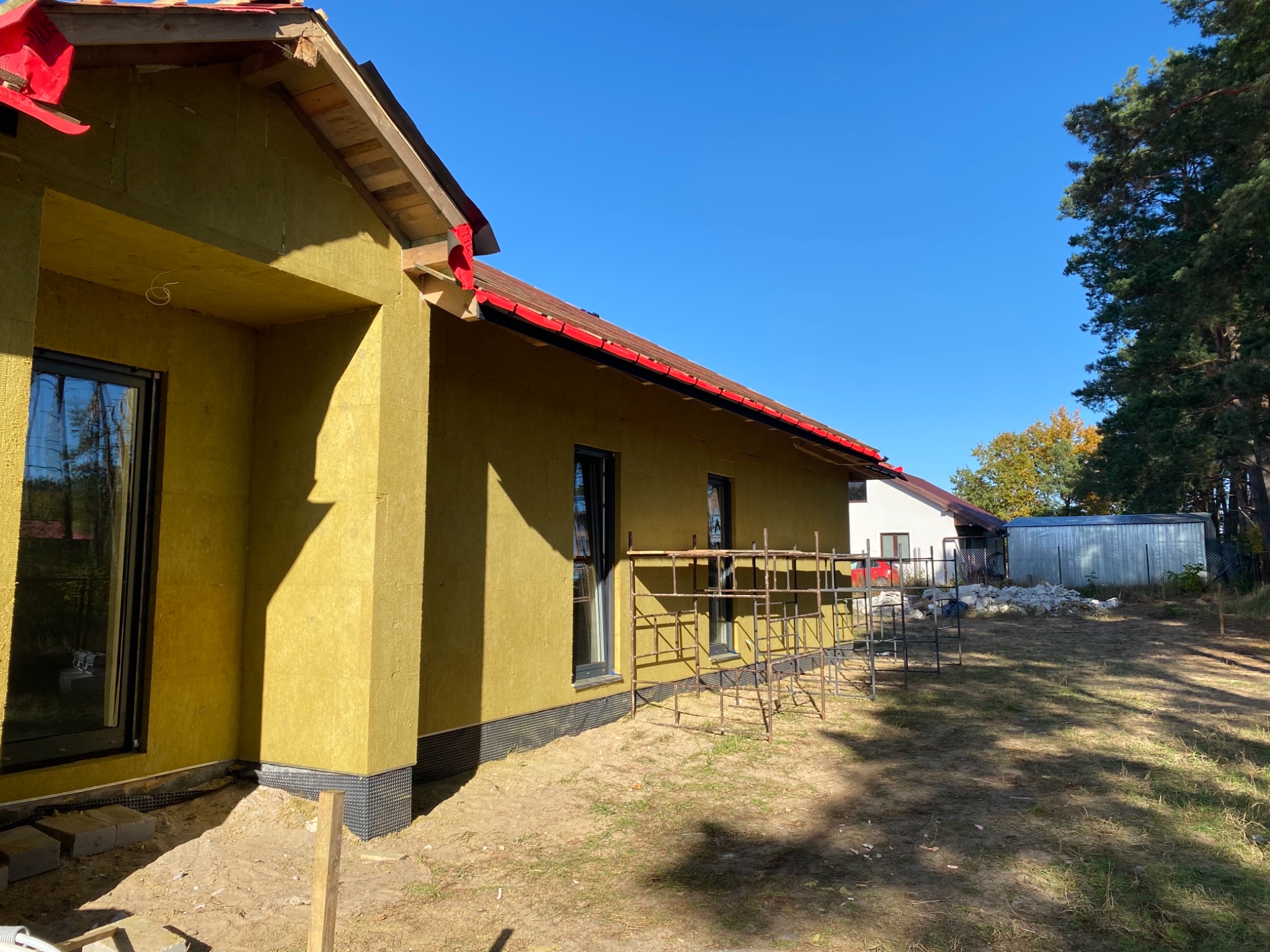 Dom jednorodzinny w trakcie ocieplania wełną mineralną, widoczne rusztowania, czarne okna, czerwona blacha na dachu, błękitne niebo w tle.