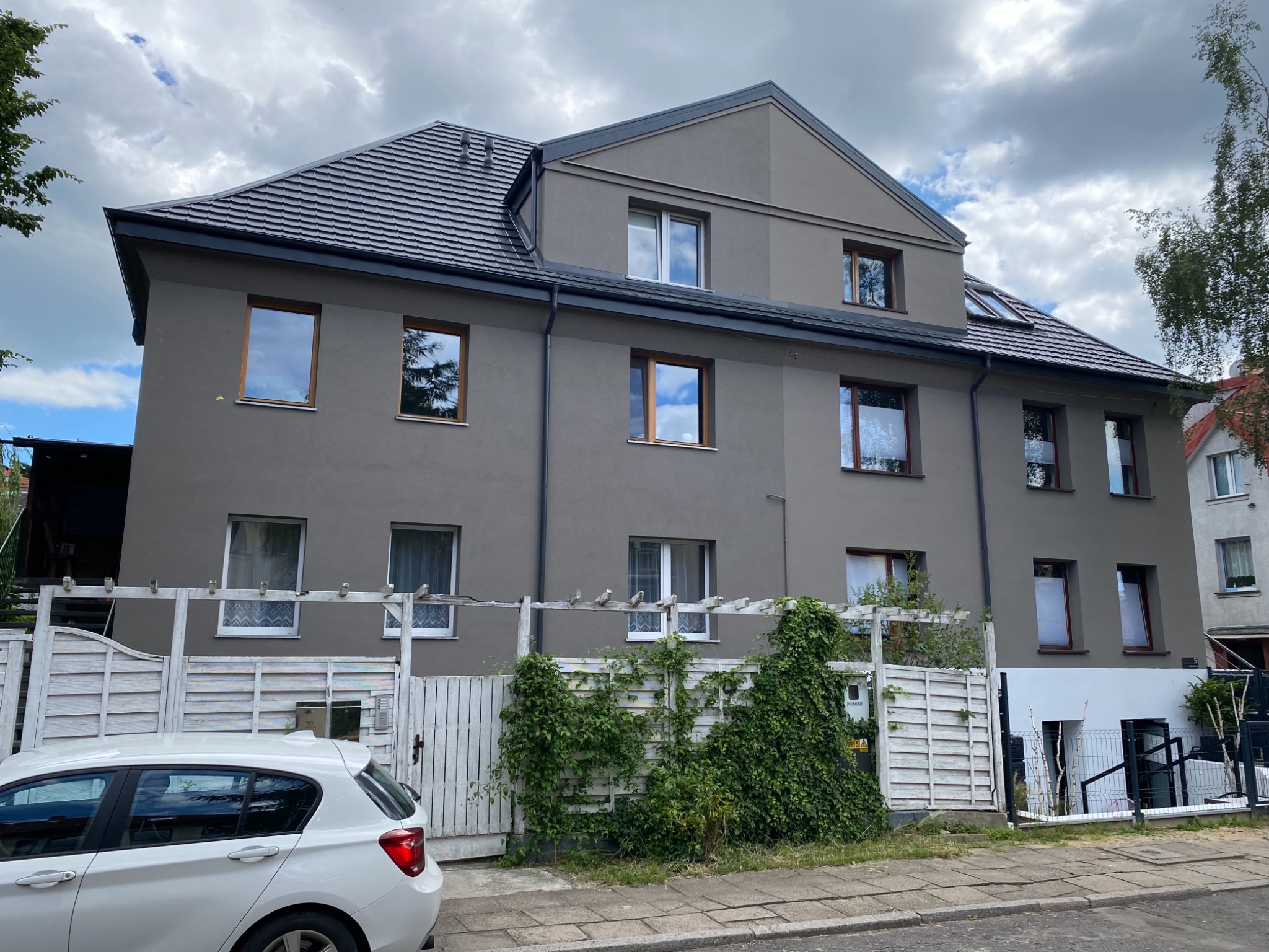 Szary, trzypiętrowy dom po kompleksowym ociepleniu, z ciemną dachówką, drewnianymi oknami i białym ogrodzeniem porośniętym zielonym bluszczem, częściowo zasłonięty przez biały samochód.