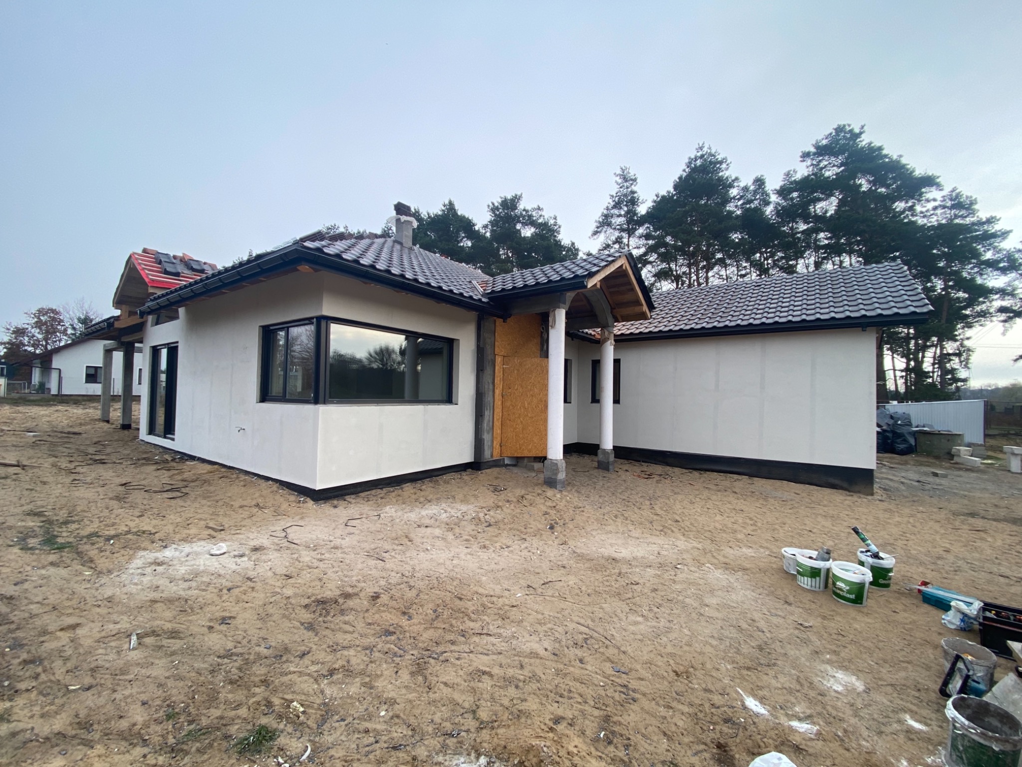 Dom jednorodzinny w trakcie budowy z otynkowaną elewacją, widoczne puszki z farbą, narzędzia i fragmenty rusztowania, drzewa w tle.