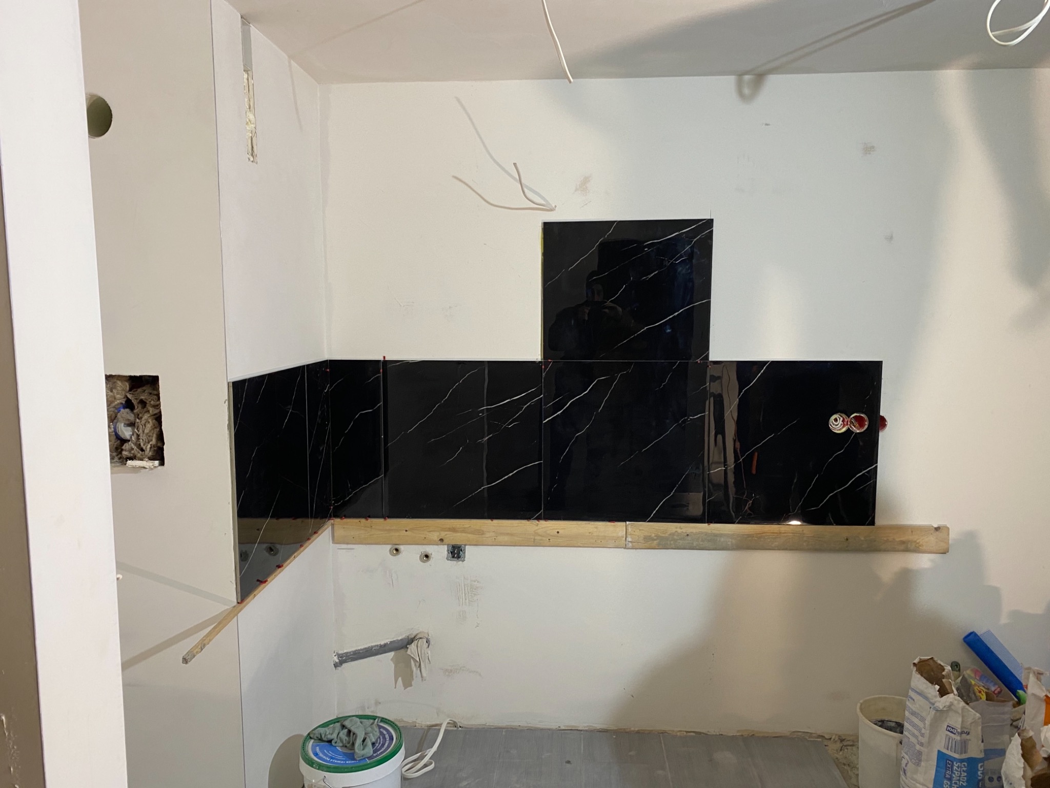 Częściowo ułożone czarne płytki z białym żyłowaniem na ścianie w trakcie remontu kuchni, widoczne drewniane listwy podtrzymujące, puszki elektryczne i akcesoria glazurnicze.