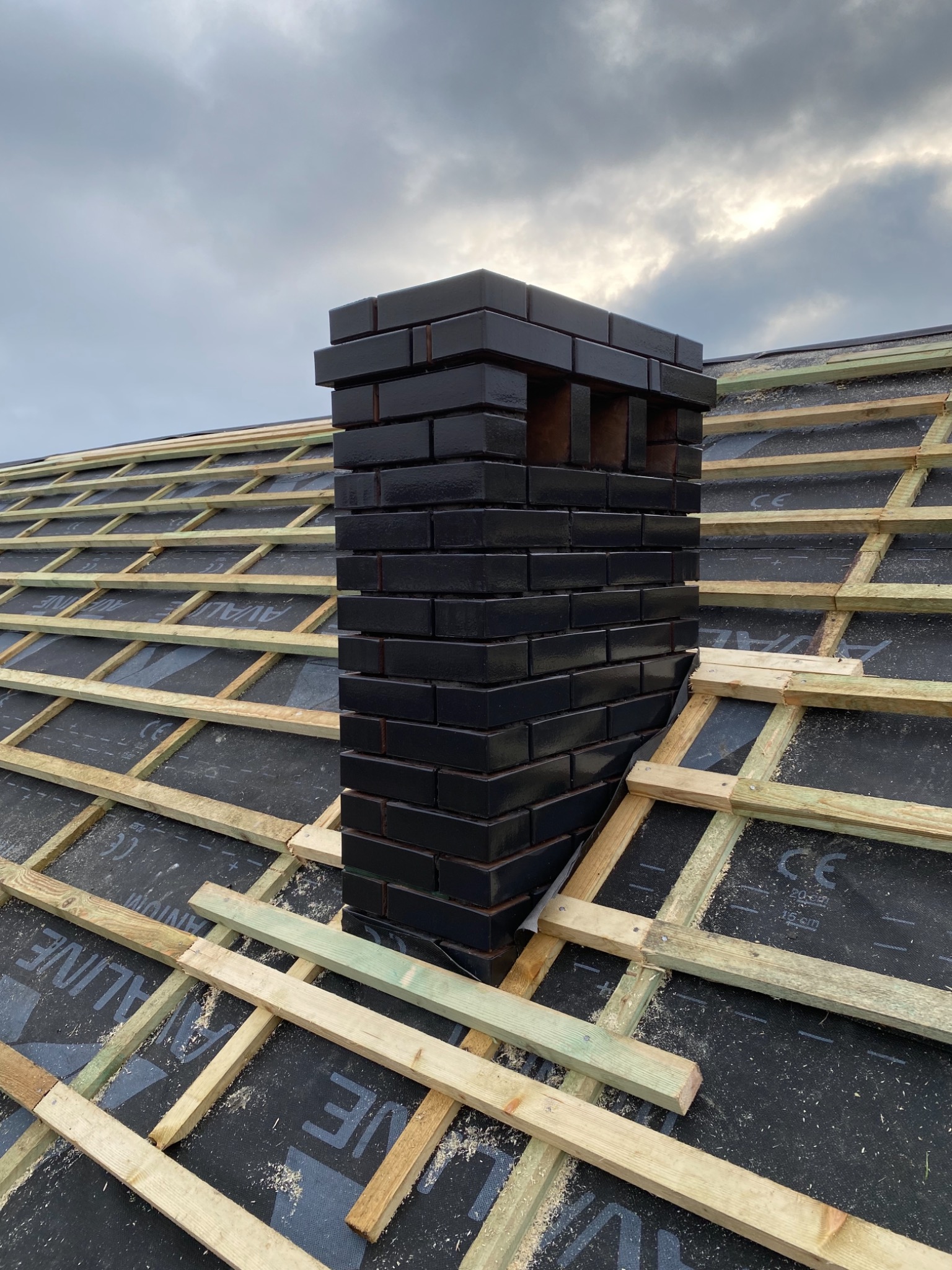 Czarny komin z cegły klinkierowej na dachu w trakcie budowy, widoczne drewniane łaty i membrana dachowa, pochmurne niebo w tle.