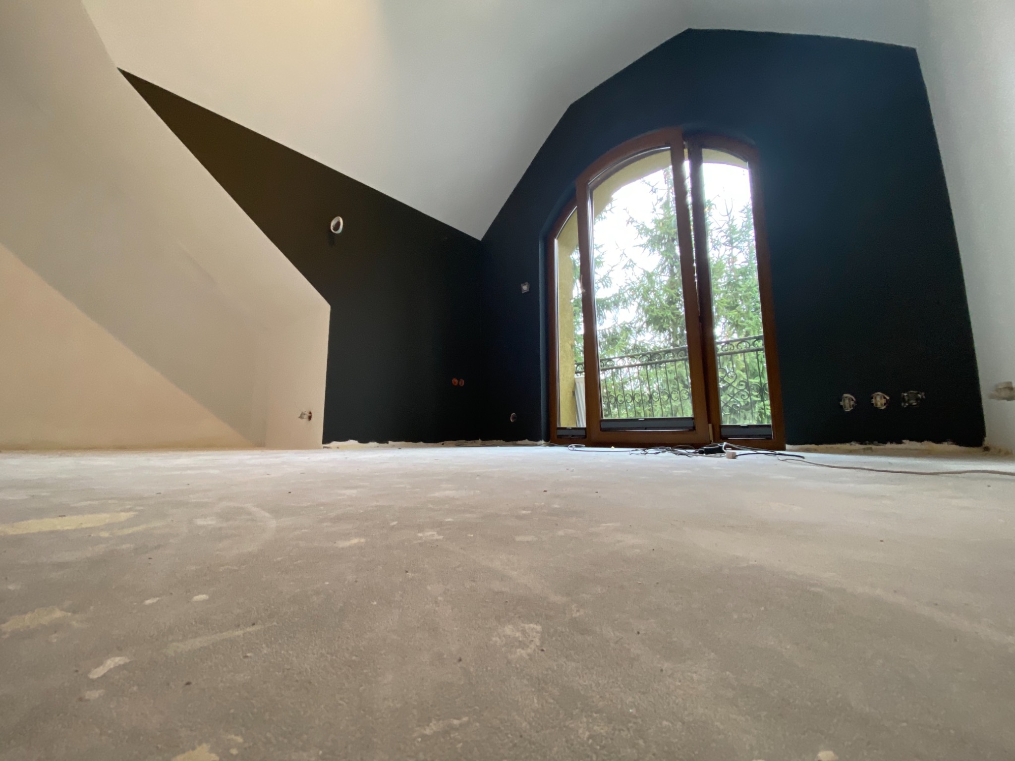 Pokój na poddaszu po malowaniu ścian na ciemny kolor, widoczne przygotowane podłoże pod podłogę i okno balkonowe z widokiem na drzewa.