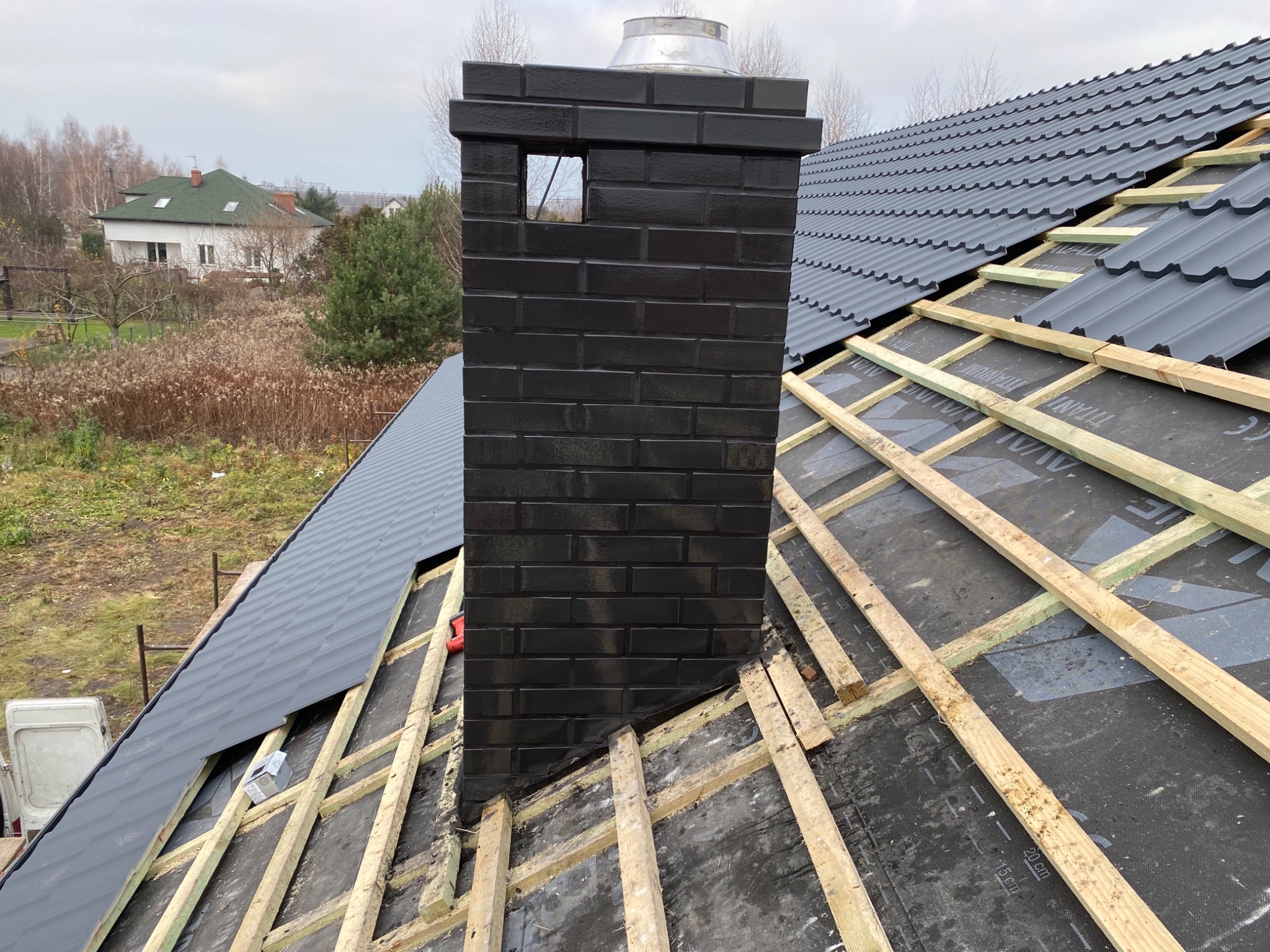 Montaż grafitowej dachówki na drewnianej konstrukcji z widocznym czarnym, ceglanym kominem z otworem wentylacyjnym i fragmentem krajobrazu w tle.